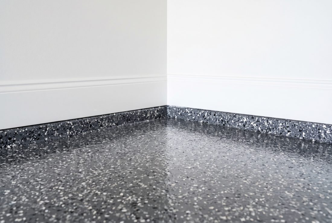 Corner of a finished charcoal and white flake floor meeting a white wall, the flake carried tight to the wall line with no bare concrete showing